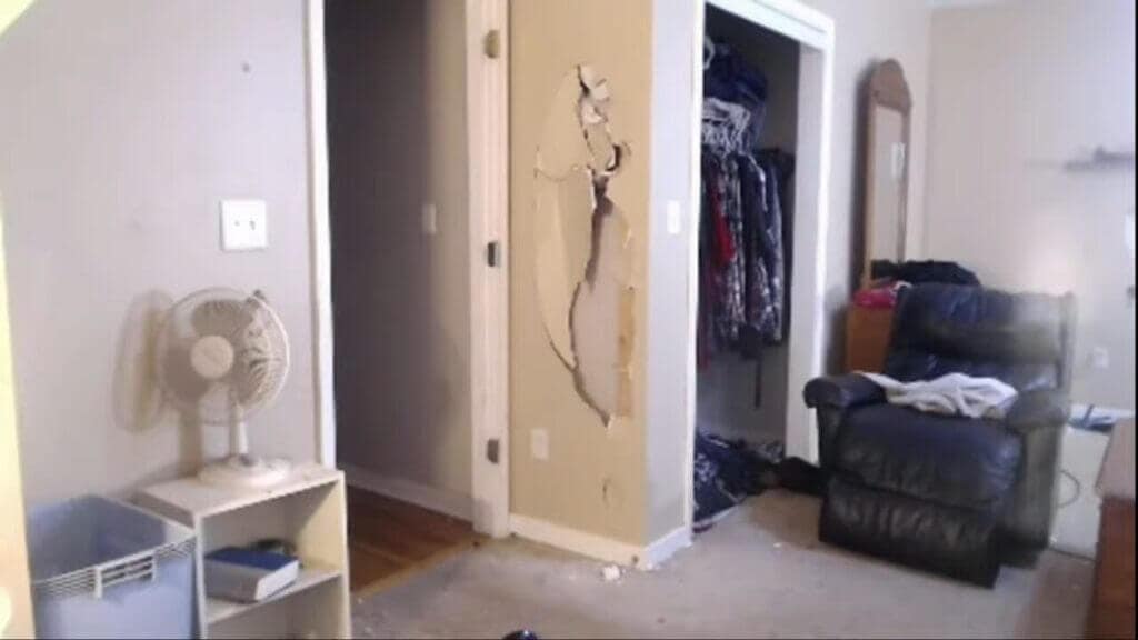BossmanJack's bedroom showing extensive damage including a destroyed door with visible cracks and holes, damaged walls, a small fan on a white shelf, and a dark chair with clothes